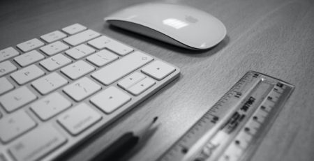 black and white image of a computer keyboard, mouse, ruler, and pencil sitting on a desk
