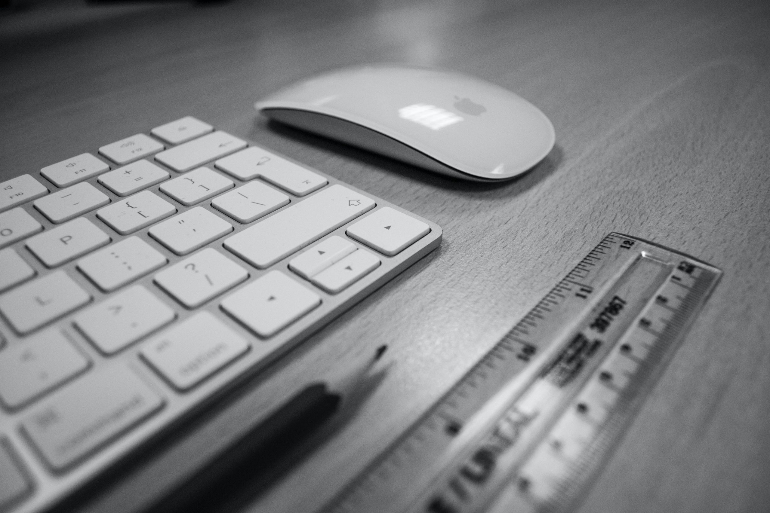 black and white image of a computer keyboard, mouse, ruler, and pencil sitting on a desk